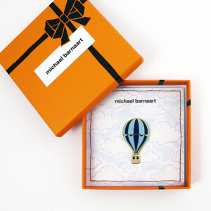Broche in de vorm van een luchtballon, in de kleuren blauw en goud. De broche zit in een oranje cadeaudoosje